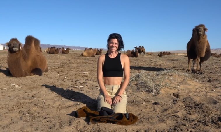 A person kneels and smiles in a desert setting, surrounded by several relaxed camels under a clear blue sky